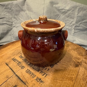 以下が含まれることがあります： 蓋付きの、ヴィンテージの釉薬をかけたセラミック製の豆ポット。ポットは濃い茶色で、縁は明るいタン色、蓋には小さなハンドルが付いています。両側に2つのハンドルがあり、光沢のある仕上がりです。