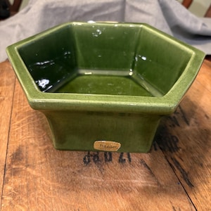 Op de afbeelding: Een groene, zeshoekige keramische plantenbak met een glanzende afwerking. De plantenbak heeft een rechthoekige basis en een licht uitlopende bovenkant. Een kleine, rechthoekige sticker is aan de zijkant bevestigd. De plantenbak staat op een houten oppervlak.