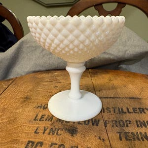 May include: A white, vintage-style glass compote dish. The bowl has a diamond pattern and a scalloped rim. It sits on a pedestal base with a round foot. The dish is displayed on a wooden surface.