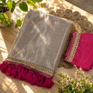 Op de afbeelding: Een glanzende, lichtgrijze stof met een decoratieve rand van gouden afwerking en magenta kwasten. Er is ook een vierkante magenta stof met gouden afwerking. De items worden gepresenteerd op een geweven mat.