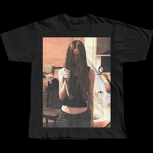 Könnte beinhalten: Schwarzes T-Shirt mit einem Bild im Vintage-Stil einer Person, die ein Messer hält. Das Bild hat einen Sepia-Ton, die Person trägt ein dunkles Oberteil und Jeans. Der Hintergrund deutet auf eine Küche hin.
