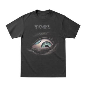 May include: Black t-shirt featuring the word "TOOL" in silver lettering above a detailed eye graphic. The eye has blue and green irises, with intricate details. The shirt is a classic crew neck style.