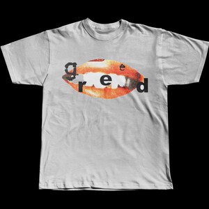 Könnte beinhalten: Hellgraues T-Shirt mit einer Mundgrafik und dem Wort "greed". Der Mund ist orange und rot, mit weißen Zähnen. Das Wort "greed" ist in Schwarz, mit einer verwaschenen Schrift.