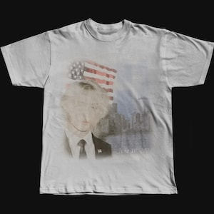 May include: A light gray t-shirt featuring a watercolor-style portrait of a person with an American flag and a cityscape in the background. The text "June 10, 20" is printed below the portrait.
