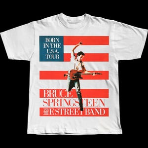 May include: White t-shirt featuring a graphic of Bruce Springsteen playing guitar, set against a red and white flag design. The shirt includes the text "BORN IN THE U.S.A. TOUR" and "BRUCE SPRINGSTEEN AND THE STREET BAND".
