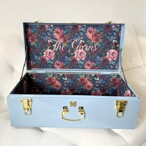 May include: Open, light blue metal trunk with gold-colored hardware. The interior is lined with a floral pattern of pink and red roses on a dark blue background. The text "The Goans" is displayed inside the lid.