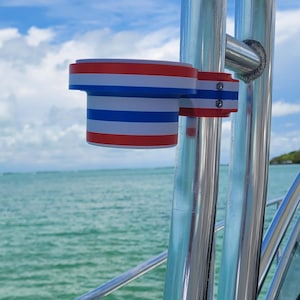 May include: A cylindrical cup holder mounted to a vertical metal pole on a boat. The holder features horizontal stripes in red, white, and blue. A second, light blue cup holder is visible further down the same metal pole, which is set against a sea.