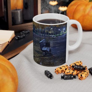 May include: White ceramic mug with a park scene printed on it, featuring a bench, trees, and a pathway. The mug is filled with a dark liquid, possibly coffee or tea. Halloween-themed candies and a pumpkin are nearby.