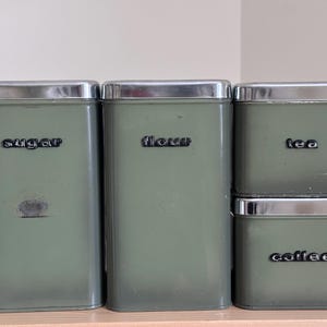 Puede incluir: Cuatro botes de cocina vintage, etiquetados como "azúcar", "harina", "té" y "café". Los botes son de color verde apagado con tapas plateadas y letras negras. Están colocados sobre una superficie de madera.