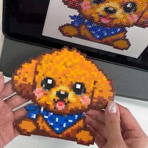 Puede incluir: Un perro pixelado hecho con cuentas naranjas, marrones y azules. El perro tiene un pañuelo azul y mejillas rosadas. Una imagen digital del mismo perro es visible en la pantalla de una tableta en el fondo.