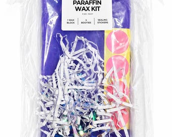 May include: A paraffin wax kit for feet in a clear plastic bag containing a large purple wax block, four white booties, and a sheet of pink circular sealing stickers. The kit includes shredded white paper filler and a white label with black text.