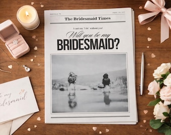 May include: A newspaper titled The Bridesmaid Times with the headline Will you be my BRIDESMAID? and a black and white photo of two people. Included are a pink ring box with a ring, a candle, a silver pen, a card, a pink ribbon, and white roses.