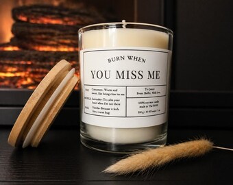 May include: A clear glass candle with white wax and a bamboo lid. The label reads: BURN WHEN YOU MISS ME, listing scent notes of cinnamon, lavender, and vanilla. It specifies 100% soy wax, made in The PNW, 200 g / 30-50 hours / 7 oz.
