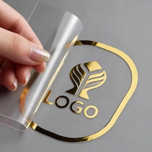 May include: A gold-colored logo with the word "LOGO" is partially revealed as a clear sticker is peeled back. The logo features a stylized design within a gold oval frame, set against a gray background.