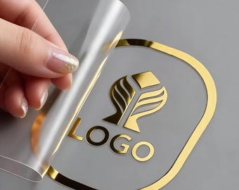 May include: A clear adhesive sticker with a metallic gold border and a central gold emblem featuring a stylised leaf design above the word LOGO in capital letters. The sticker is partially peeled back from its transparent backing layer.