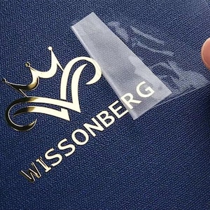 May include: A gold-coloured logo featuring a crown design and the word "WISSONBERG" on a textured blue surface. A clear adhesive strip is partially peeled back, revealing the logo's text.