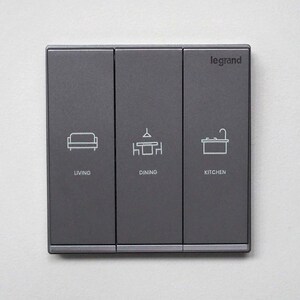 May include: A dark gray light switch panel with three sections. Each section has a white icon and text: "LIVING", "DINING", and "KITCHEN". The brand name "legrand" is printed in the upper right corner.