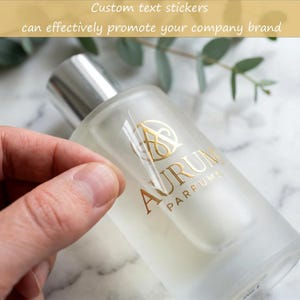 Puede incluir: Una pegatina transparente con letras de lámina dorada que dicen AURUM PARFUMS y un logotipo estilizado, aplicada a un frasco de perfume de vidrio esmerilado con tapa plateada. El texto superior dice Custom text stickers can effectively promote your company brand.