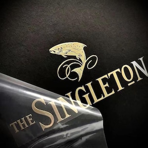 Könnte beinhalten: Goldfolienlogo mit einem aufspringenden Fisch und dem Schriftzug "The Singleton" in goldener Schrift auf schwarzem Hintergrund. Eine transparente Kunststofffolie wird teilweise abgezogen.