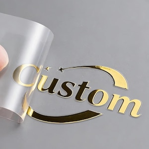 May include: Gold foil sticker with the word "Custom" in a bold, elegant font. The sticker has a star and curved design element. The sticker is partially peeled back, revealing the adhesive backing. The background is a light grey.