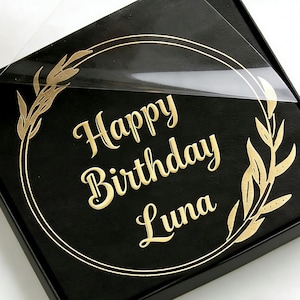 May include: A black gift box featuring the gold text "Happy Birthday Luna" within a gold wreath. The box has a clear plastic cover, partially peeled back. The design is elegant and suitable for a birthday celebration.