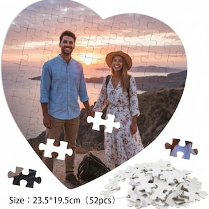 Peut inclure: Puzzle en forme de cœur avec un couple et un coucher de soleil en arrière-plan. Le puzzle mesure 23,5 x 19,5 cm (52 pièces). Des pièces de puzzle blanches sont éparpillées autour du puzzle assemblé.