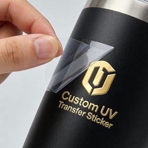 Peut inclure: Un gobelet noir mat avec un logo métallique doré et le texte Custom UV Transfer Sticker. Un film adhésif transparent est décollé de la surface, révélant le lettrage et l'emblème dorés en relief appliqués sur le récipient sombre.