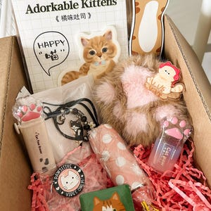 May include: A cardboard box filled with cat-themed items. Includes stickers, a plush ball, a bracelet, a wooden block, and a pink and white polka-dot item. The box contains red paper shreds and a variety of cat-themed accessories.