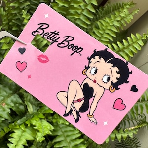 May include: A pink tag featuring the cartoon character Betty Boop. The tag has the words "Betty Boop" in black script, along with a cartoon image of Betty Boop, hearts, and a kiss mark.