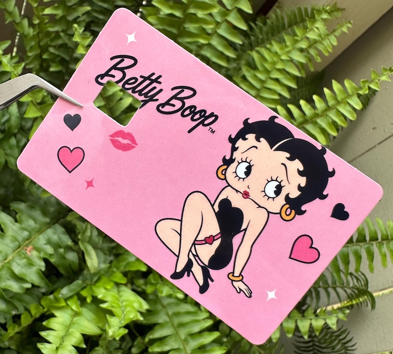 May include: A pink tag featuring the cartoon character Betty Boop. The tag has the words "Betty Boop" in black script, along with a cartoon image of Betty Boop, hearts, and a kiss mark.