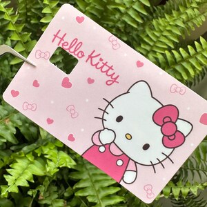 May include: A pink rectangular card featuring Hello Kitty. The card has a cut-out square and is decorated with hearts, bows, and a Hello Kitty illustration. The Hello Kitty character has a pink bow and dress.