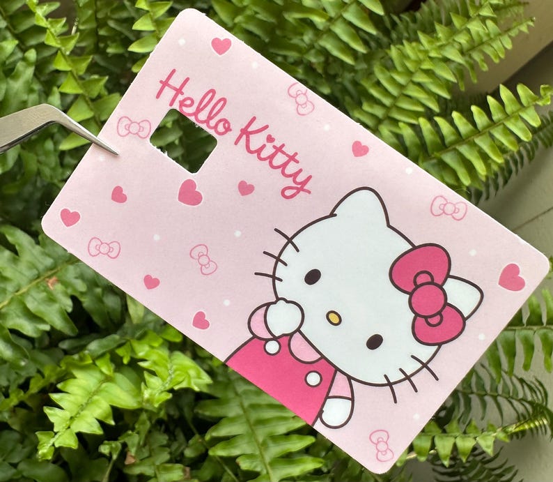 May include: A pink rectangular card featuring Hello Kitty. The card has a cut-out square and is decorated with hearts, bows, and a Hello Kitty illustration. The Hello Kitty character has a pink bow and dress.
