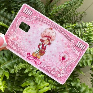 Może przedstawiać: R&oacute;żowe etui na telefon z motywem Strawberry Shortcake. Etui ma grafikę Strawberry Shortcake trzymającej koszyk, z tekstem "Strawberry Shortcake" i "One Hundred Berry Dollars". Etui ma r&oacute;wnież numer "100".