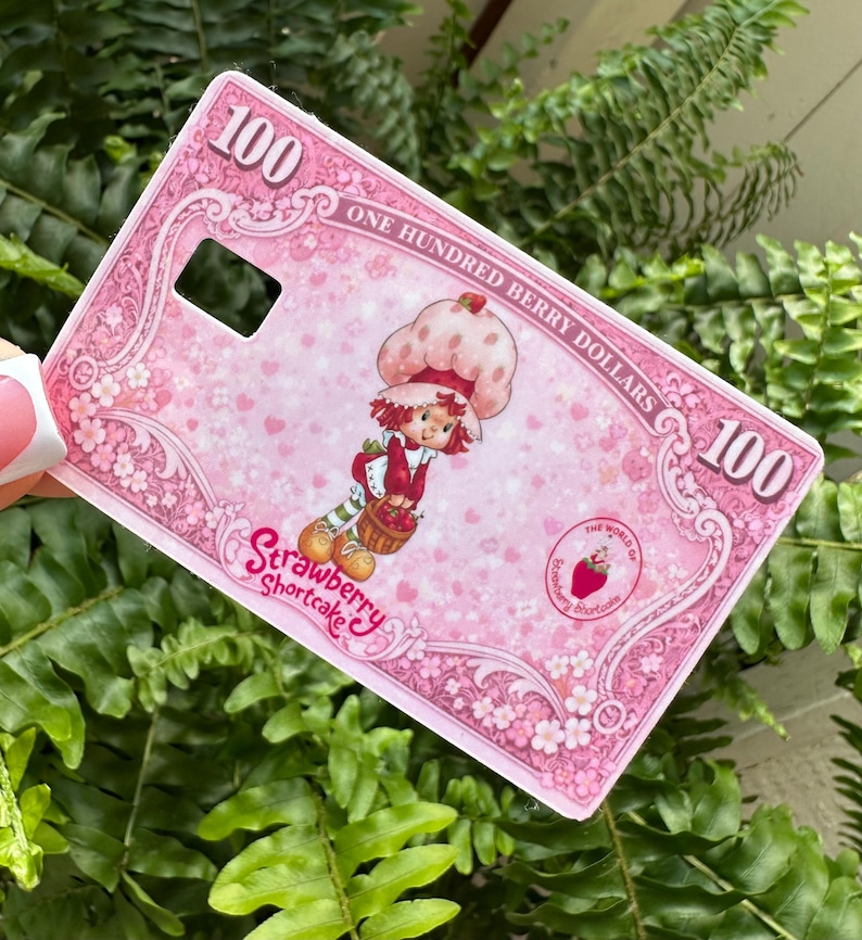 Może przedstawiać: R&oacute;żowe etui na telefon z motywem Strawberry Shortcake. Etui ma grafikę Strawberry Shortcake trzymającej koszyk, z tekstem "Strawberry Shortcake" i "One Hundred Berry Dollars". Etui ma r&oacute;wnież numer "100".