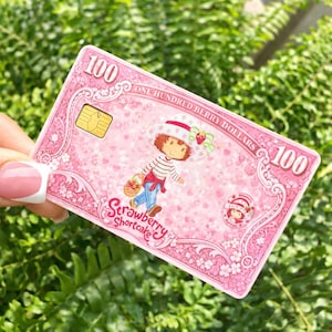 May include: A pink Strawberry Shortcake-themed card with the text "100 ONE HUNDRED BERRY DOLLARS 100". The card features an illustration of Strawberry Shortcake, a cartoon character, and a small chip. The card is held against a background of green foliage.