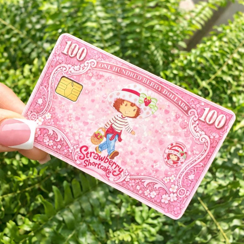 May include: A pink Strawberry Shortcake-themed card with the text "100 ONE HUNDRED BERRY DOLLARS 100". The card features an illustration of Strawberry Shortcake, a cartoon character, and a small chip. The card is held against a background of green foliage.