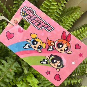 May include: A pink tag featuring the Powerpuff Girls logo and illustrations of the three characters. The tag has a heart-shaped hole and is decorated with hearts, stars, and colorful stripes.