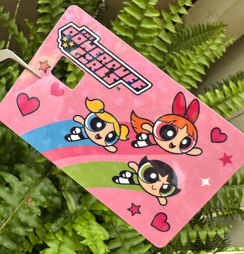 May include: A pink tag featuring the Powerpuff Girls logo and illustrations of the three characters. The tag has a heart-shaped hole and is decorated with hearts, stars, and colorful stripes.