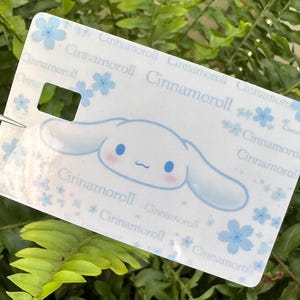 Peut inclure: Une &eacute;tiquette rectangulaire blanche avec le personnage Cinnamoroll, des accents bleus et des motifs floraux. L'&eacute;tiquette a une d&eacute;coupe rectangulaire et le texte "Cinnamoroll" r&eacute;p&eacute;t&eacute;.
