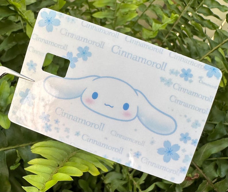 Peut inclure: Une &eacute;tiquette rectangulaire blanche avec le personnage Cinnamoroll, des accents bleus et des motifs floraux. L'&eacute;tiquette a une d&eacute;coupe rectangulaire et le texte "Cinnamoroll" r&eacute;p&eacute;t&eacute;.