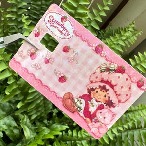 May include: A pink Strawberry Shortcake-themed tag with a cartoon illustration of Strawberry Shortcake and her cat. The tag has a pink gingham pattern and strawberry accents. The text "Strawberry Shortcake" is at the top.