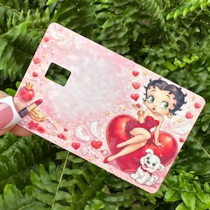 Puede incluir: Una tarjeta rosa con Betty Boop sentada en un coraz&oacute;n rojo, con un cachorro blanco en la parte inferior. La tarjeta tiene un recorte rectangular y est&aacute; decorada con corazones, perfume y l&aacute;piz labial. El fondo es rosa claro.