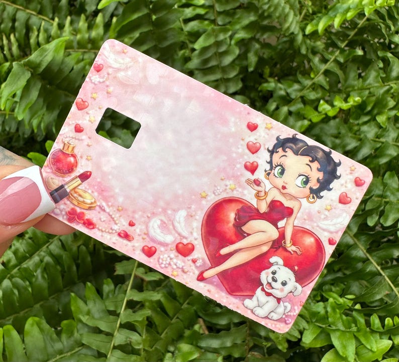 Puede incluir: Una tarjeta rosa con Betty Boop sentada en un coraz&oacute;n rojo, con un cachorro blanco en la parte inferior. La tarjeta tiene un recorte rectangular y est&aacute; decorada con corazones, perfume y l&aacute;piz labial. El fondo es rosa claro.