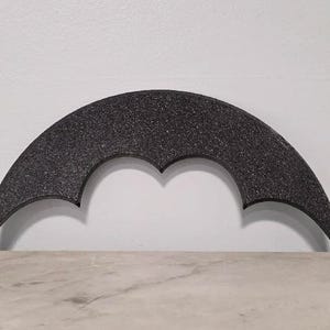 May include: A black, crescent-shaped decorative object with a textured surface. The object has a bat-like design with pointed ends and a central indentation. It is set against a white wall and a light-colored surface.