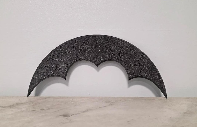 May include: A black, crescent-shaped decorative object with a textured surface. The object has a bat-like design with pointed ends and a central indentation. It is set against a white wall and a light-colored surface.