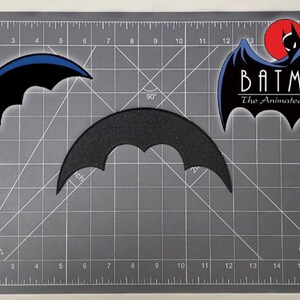May include: Three Batman-themed patches on a gray cutting mat. One patch is a black bat symbol with a blue outline, another is a textured black bat symbol, and the third features the Batman logo from "The Animated Series."