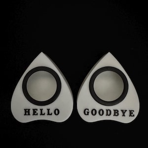 May include: Two white planchette pointers with black accents and the words "HELLO" and "GOODBYE". The pointers are teardrop-shaped with a circular opening in the center. The background is black.