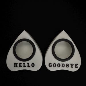 May include: Two white planchette pointers with black accents and the words "HELLO" and "GOODBYE". The pointers are teardrop-shaped with a circular opening in the center. The background is black.