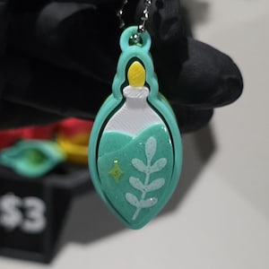 May include: A teal pendant in the shape of a potion bottle, with a white top and a yellow stopper. The bottle contains a teal liquid with a white leaf design and a yellow star. The pendant hangs from a silver chain.
