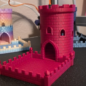 May include: A bright magenta 3D printed dice tower featuring a cylindrical castle turret with a brick texture, arched windows, and a crenelated top. The structure includes an attached square courtyard base with a low wall and small decorative spheres.